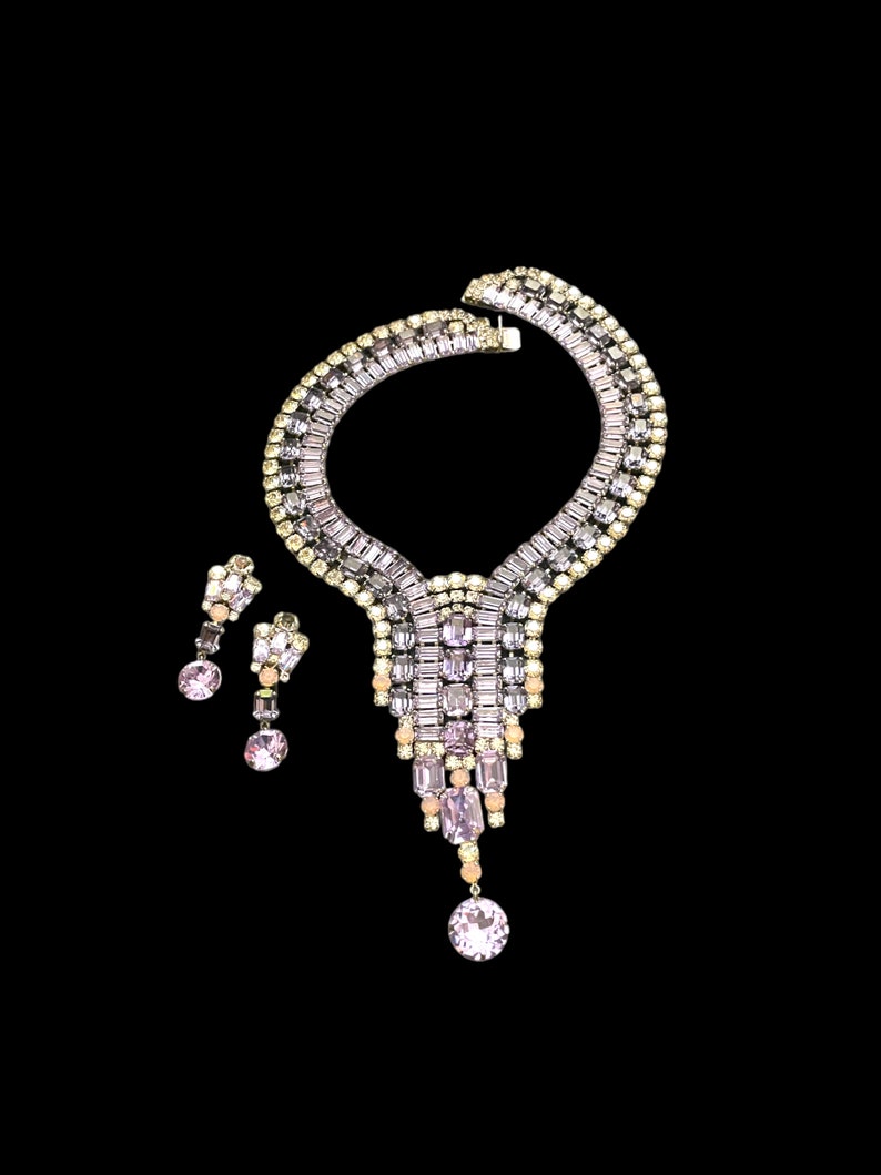 May include: A vintage rhinestone necklace and earrings set. The necklace features a large, teardrop-shaped pendant with a cascading design of clear and pink rhinestones. The earrings are simple and elegant, with a single, round rhinestone.