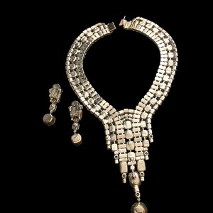 May include: A silver tone rhinestone necklace and earrings set. The necklace features a cascading design with multiple rows of rhinestones. The earrings are dangle style with a single large rhinestone.