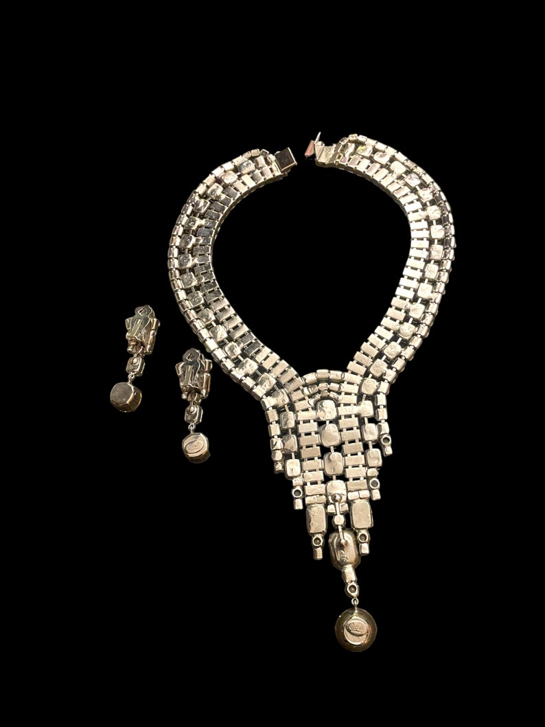May include: A silver tone rhinestone necklace and earrings set. The necklace features a cascading design with multiple rows of rhinestones. The earrings are dangle style with a single large rhinestone.