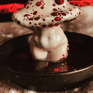 May include: A white ceramic mushroom figurine with red polka dots, sitting in a black bowl with red speckles. The mushroom has a human-like body and is a decorative object.