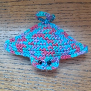 May include: A handmade crochet stingray in shades of blue, purple, and red. The aquatic animal has small black eyes and a loop for hanging. The stingray is made with a textured stitch.