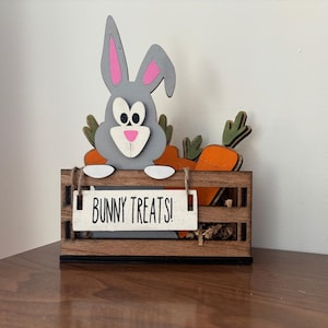 May include: A wooden crate with a grey bunny rabbit peeking out of the top. The crate is filled with brown paper shreds and has a sign that says "Bunny Treats!"