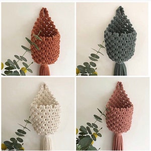 May include: Four macrame wall hanging planters in different colours: terracotta, grey, white, and pink. Each planter has a tassel at the bottom.
