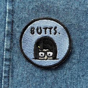 May include: A blue and black embroidered patch with the text "BUTTS." and a cartoon image of a woman with black hair and glasses.