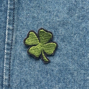 May include: A green embroidered shamrock patch with a black border is centered on a blue denim background. The four-leaf clover design is a symbol of good luck and is commonly associated with St. Patrick's Day.