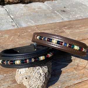 May include: Two leather dog collars with beaded trim. The top collar is brown leather with a beaded trim of dark blue, green, white, and gold beads. The bottom collar is black leather with a beaded trim of dark blue, red, white, and gold beads.