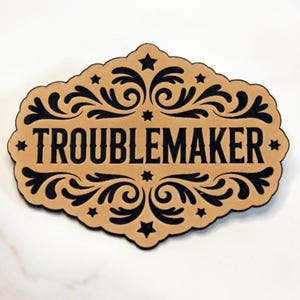 May include: A tan-colored decorative plaque with the word "TROUBLEMAKER" in bold black letters. The plaque has a decorative black scroll design and star accents. The background is white.
