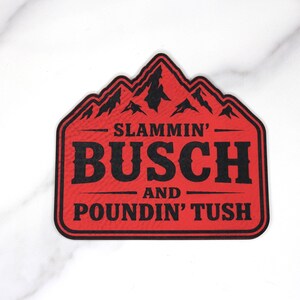 May include: A red and black sticker featuring the text "SLAMMIN' BUSCH AND POUNDIN' TUSH" within a mountain-shaped design. The sticker is rectangular with a mountain range at the top.