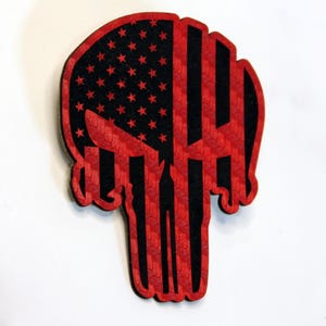 Op de afbeelding: Een rood en zwart schedelvormig embleem met een verweerd Amerikaans vlagontwerp. Het bovenste gedeelte heeft een zwart veld met rode sterren, terwijl de onderste helft rode en zwarte verticale strepen heeft.
