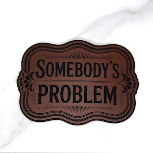May include: A brown sign with the words "SOMEBODY'S PROBLEM" in black, framed by a decorative border. The sign has a vintage aesthetic and is set against a white background.