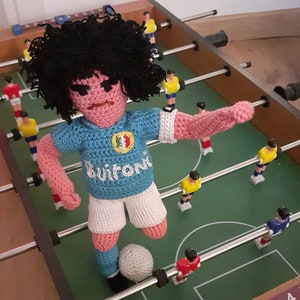 Puede incluir: Mu&ntilde;eco de jugador de futbol&iacute;n hecho a mano con ganchillo, con pelo negro, que lleva una camiseta azul claro con un logotipo blanco y la palabra "Buitoni". El mu&ntilde;eco est&aacute; colocado en una mesa de futbol&iacute;n verde, listo para chutar un bal&oacute;n blanco.