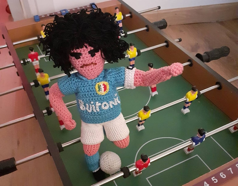 Puede incluir: Mu&ntilde;eco de jugador de futbol&iacute;n hecho a mano con ganchillo, con pelo negro, que lleva una camiseta azul claro con un logotipo blanco y la palabra "Buitoni". El mu&ntilde;eco est&aacute; colocado en una mesa de futbol&iacute;n verde, listo para chutar un bal&oacute;n blanco.