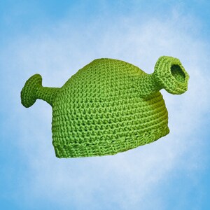 Pode incluir: Chapéu de crochê verde em forma de ogro de desenho animado. O chapéu tem uma orelha grande de um lado e uma orelha menor do outro.