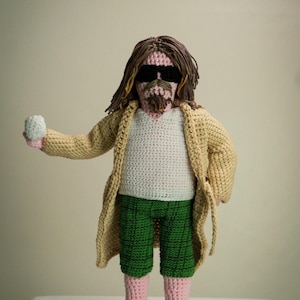 Può includere: Bambola all'uncinetto che indossa una vestaglia color sabbia, una camicia bianca, pantaloncini verdi e sandali marroni. La bambola ha i capelli castani, il viso rosa e occhiali da sole neri.