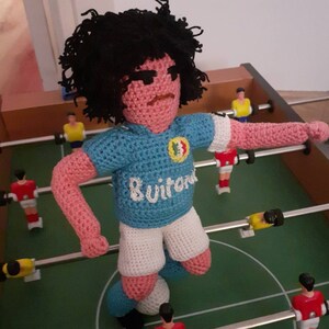Puede incluir: Mu&ntilde;eco de ganchillo que representa a un jugador de f&uacute;tbol con camiseta azul claro y pantal&oacute;n corto blanco. El mu&ntilde;eco, con pelo negro, est&aacute; sobre un futbol&iacute;n, con otros jugadores visibles. La camiseta lleva la palabra "Suitor".