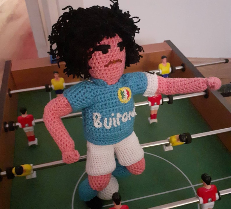 Puede incluir: Mu&ntilde;eco de ganchillo que representa a un jugador de f&uacute;tbol con camiseta azul claro y pantal&oacute;n corto blanco. El mu&ntilde;eco, con pelo negro, est&aacute; sobre un futbol&iacute;n, con otros jugadores visibles. La camiseta lleva la palabra "Suitor".