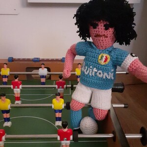 Puede incluir: Mu&ntilde;eco de futbolista de ganchillo con pelo negro, que lleva una camiseta azul claro con la palabra "Vitoni" y pantalones cortos blancos. El mu&ntilde;eco est&aacute; en un futbol&iacute;n, con jugadores en miniatura rojos y amarillos.