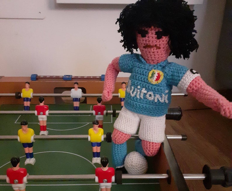 Puede incluir: Mu&ntilde;eco de futbolista de ganchillo con pelo negro, que lleva una camiseta azul claro con la palabra "Vitoni" y pantalones cortos blancos. El mu&ntilde;eco est&aacute; en un futbol&iacute;n, con jugadores en miniatura rojos y amarillos.