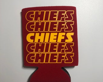 May include: A maroon can cooler with the word "CHIEFS" printed in yellow multiple times. The cooler has a cutout at the bottom for easy gripping and is designed to keep beverages cold. The background is a plain white surface.