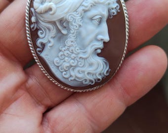 May include: Oval cameo pendant featuring a profile of a bearded man in white relief against a brown backdrop. The pendant has a silver-coloured frame and a loop for a chain.