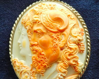 May include: A circular pendant with a cameo design. It features two profiles in relief, with the central figure having orange hair and a beard. The pendant has a gold-coloured frame and bail. The cameo is set against a cream background.
