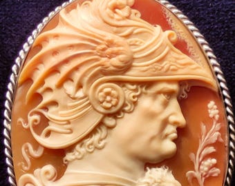 May include: Oval cameo pendant featuring a profile of a figure in a helmet, carved in cream and orange hues. The pendant has a silver-coloured frame and bail. The cameo displays detailed carvings, including a lion and floral designs.