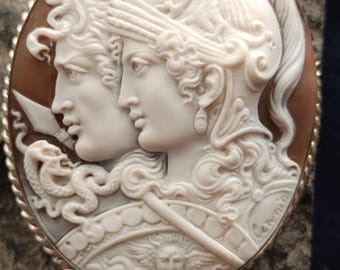 May include: Oval cameo pendant featuring two classical figures in profile, carved in white against a brown background. The figures are adorned with detailed hairstyles and armour, set in a silver-toned frame.