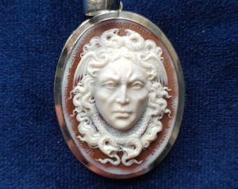 May include: Oval pendant featuring a cameo of a classical figure. The white cameo is set against a brown background, framed by a silver-coloured metal. The figure has detailed facial features and curly hair.