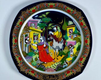 May include: A square porcelain decorative plate featuring a colourful illustration of a figure in a red robe and a person on a horse amidst buildings and trees. The border is dark blue with intricate gold, red, and green patterns. Signed Bjorn Wiinblad.
