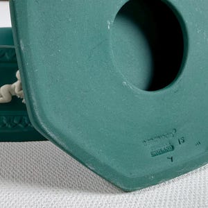 May include: A teal ceramic candle holder with a cherub relief. The candle holder has a square base with a decorative pattern and a circular opening. The underside of the lid has the text "ENGLAND".