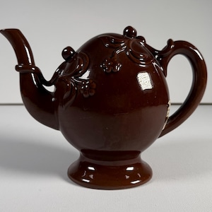 May include: A glossy dark brown ceramic teapot featuring a rounded body, a curved spout, and a handle. The exterior surface is decorated with raised, molded floral and vine relief patterns that wrap around the upper portion of the teapot.