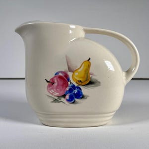 May include: Cream-colored ceramic pitcher with a curved handle and a spout. The pitcher is decorated with a colorful illustration of a yellow pear, red apple, purple plums, and blue grapes. The pitcher has a ribbed base.