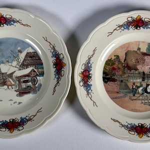 Puede incluir: Dos platos decorativos con bordes festoneados. Cada plato presenta una escena central de un pueblo, una en invierno y otra con un carruaje tirado por caballos. Diseños florales y de vid en rojo, azul y amarillo adornan los bordes.