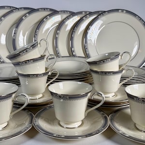 Op de afbeelding: Een complete crèmekleurige servies set met een marineblauwe en zilveren rand. De set bevat borden, schotels en theekopjes. De theekopjes hebben een gebogen handvat. Ideaal voor formele diners.