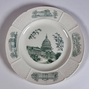 May include: A white ceramic plate featuring a dark green transfer print of the United States Capitol building in the centre. The rim is decorated with four smaller illustrations of government buildings and four sections of a raised basketweave texture.