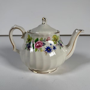 Puede incluir: Una tetera de porcelana color crema con cuerpo acanalado y tapa a juego. La tetera presenta un patrón floral alrededor del centro, mostrando rosas rosadas, flores azules y hojas verdes, con detalles en oro en el asa y el pico vertedor.