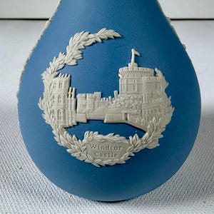 Może przedstawiać: Matowe niebieskie naczynie ceramiczne z wypukłym białym reliefem przedstawiającym zamek Windsor. Zamek otoczony jest szczegółowym wieńcem laurowym, a u podstawy okrągłego motywu dekoracyjnego znajduje się biały napis Windsor Castle.