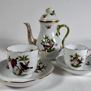 May include: A white porcelain tea set featuring a coffee pot, two teacups, and saucers. The set is decorated with colorful bird and branch illustrations, gold trim, and a green handle. The coffee pot has a silver spout.