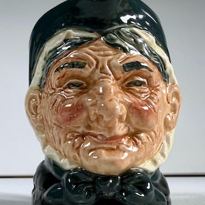 May include: A ceramic figurine of an elderly person's head, featuring a detailed face with wrinkles, rosy cheeks, and a smiling expression. The figure wears a dark green hat, a white collar, and a dark green bow tie. The figurine is a collectible item.