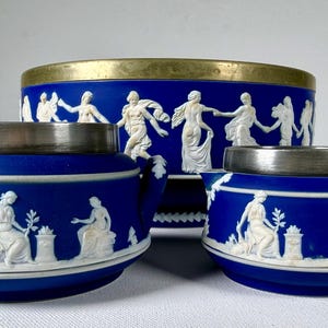 May include: A set of three blue ceramic vessels with white relief figures. The large bowl has a brass rim, while the two smaller lidded containers feature silver-toned metal rims and handles. Each piece displays classical scenes of figures.