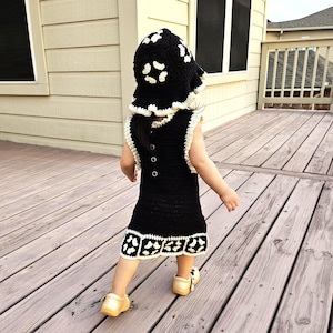 Könnte beinhalten: Ein schwarzes Häkelkleid und ein passender Hut mit weißen Verzierungen und Akzenten. Das Kleid hat einen Knopfverschluss hinten und eine dekorative Bordüre. Der Hut hat eine breite Krempe und ist mit weißen Blumenformen verziert. Das Outfit wird mit gelben Schuhen kombiniert.