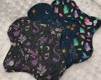 May include: Three reusable cloth menstrual pads with different patterns. The first pad has a black background with a purple, pink, and white design of crystals, eyes, and snakes. The second pad has a dark blue background with a gold star and moon design. The third pad has a black background with a green, blue, and purple crystal design.