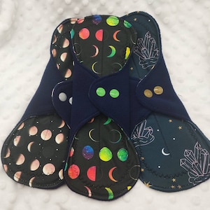 May include: Three reusable cloth menstrual pads with different patterns. The first pad has a dark background with a white moon and stars pattern. The second pad has a black background with a colorful moon and stars pattern. The third pad has a dark blue background with a white crystal pattern.