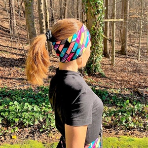 May include: A woman with a ponytail wears a black shirt and colorful patterned headband. She is standing in a wooded area.