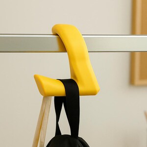 May include: A bright yellow hook is attached to a silver bar. A black strap secures a black bag to the hook. The hook has a modern, minimalist design, suitable for hanging items.
