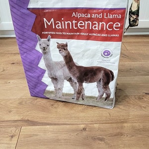Puede incluir: Una bolsa blanca con un diseño morado y blanco. La bolsa tiene el texto "Mazuri Alpaca and Llama Maintenance Fortified Feed to Maintain Adult Alpacas and Llamas" impreso en ella. La bolsa también tiene una imagen de dos alpacas.