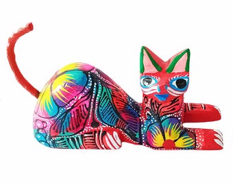 Può includere: Statuetta di gatto in legno dipinta a mano in posizione reclinata. Il gatto è principalmente rosso con motivi intricati in colori vivaci tra cui rosa, blu, giallo e verde. La coda è arricciata e gli occhi sono blu.