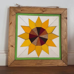 May include: Wooden framed wall art featuring a sunflower design. The sunflower has yellow petals and a center of various brown shades. The artwork is set against a white background with a green border, all within a natural wood frame.
