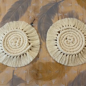 Peut inclure: Deux dessous-de-verre en crochet blanc avec des franges. Les dessous-de-verre sont ronds et présentent un motif circulaire au centre.
