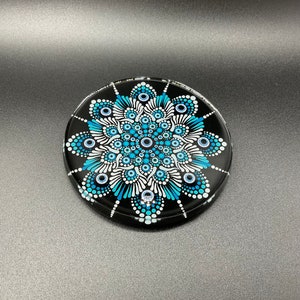 Op de afbeelding: Een ronde zwarte onderzetter met een blauw en wit mandala-ontwerp. De mandala heeft een cirkelvormig patroon van stippen en spiralen.